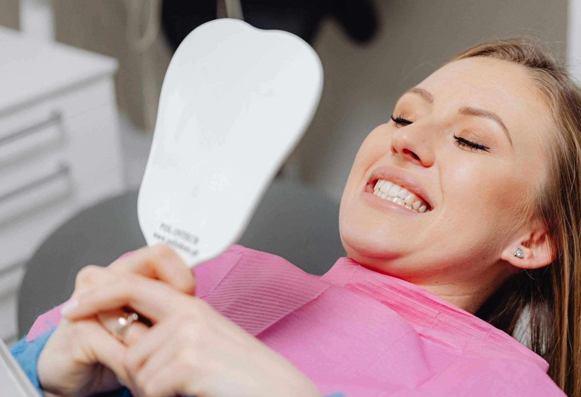 A woman at a dental clinic smiles while holding a mirror, showcasing her dental treatment results.