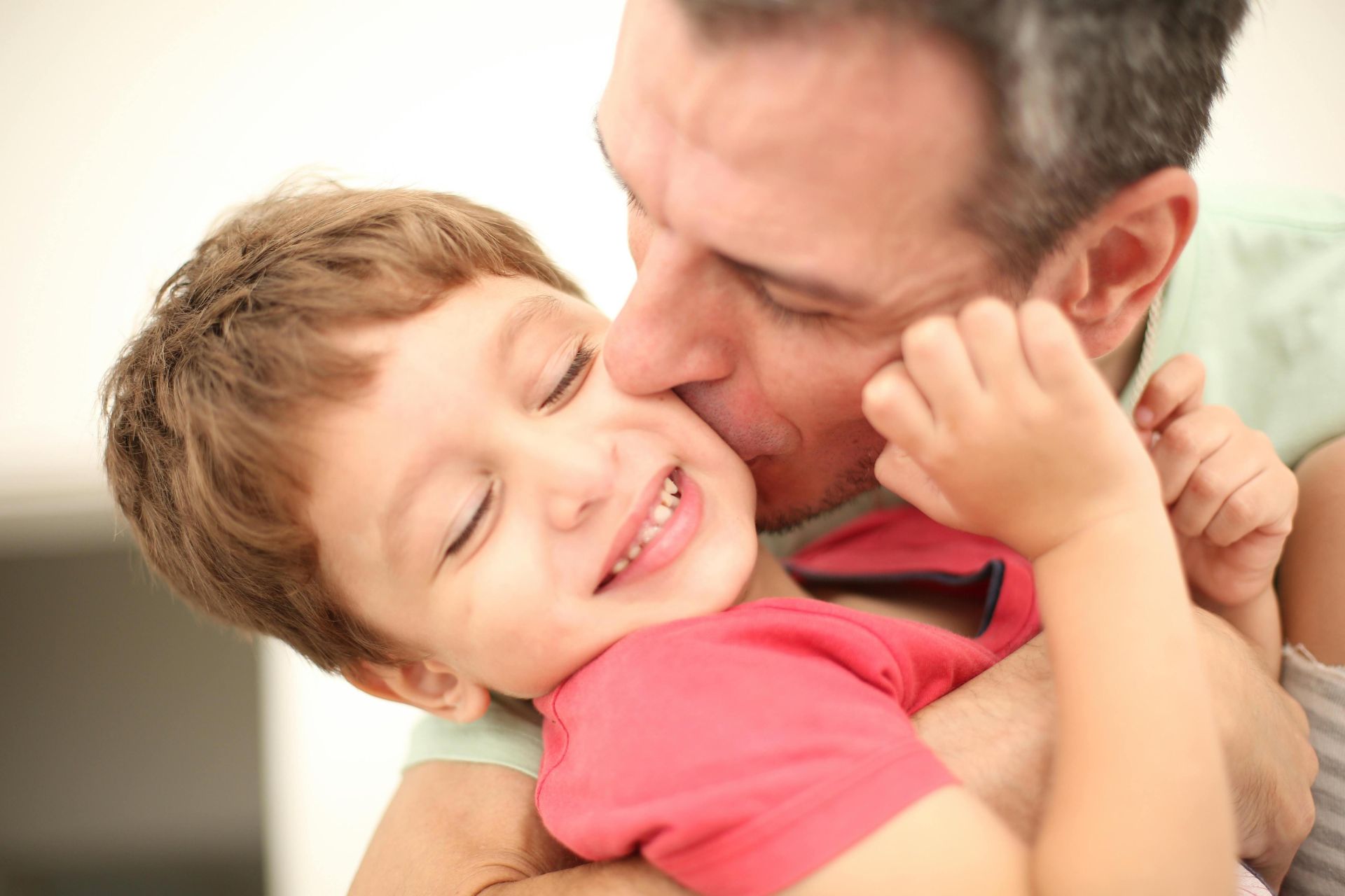 A joyful father affectionately kisses his smiling son indoors, capturing a moment of family love and happiness.